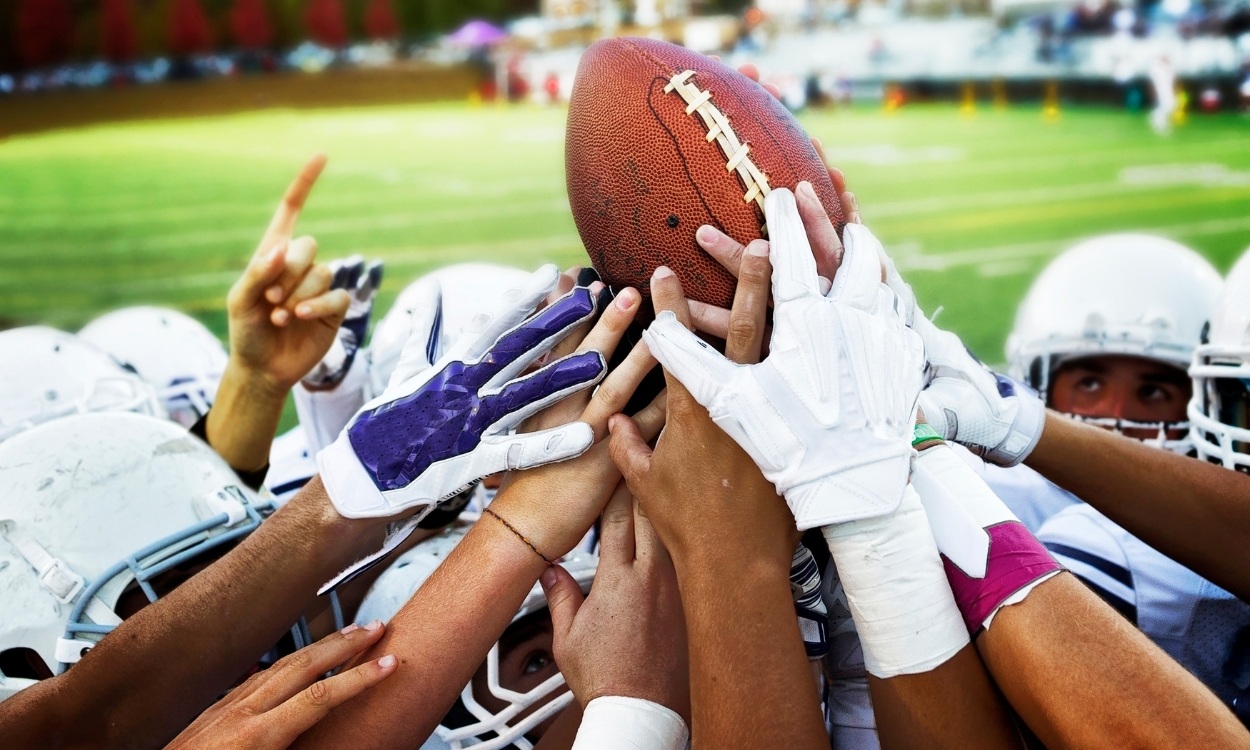 football team with all hands gathered in center holding football - teamwork and victory concept