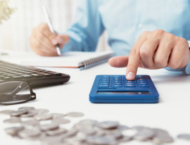 A person is using a calculator with their right hand and writing with a pen in their left hand. There is a keyboard, a notebook, glasses, and a pile of coins on the table.